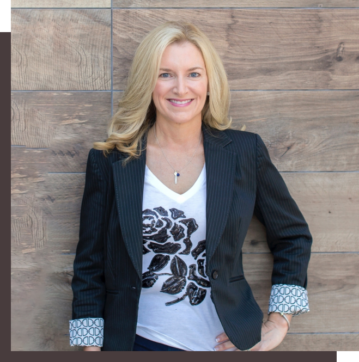 Smiling woman in a black blazer and white top poses against a wooden background.