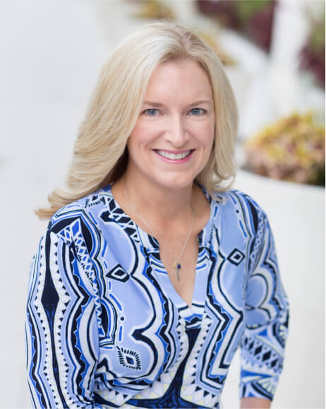Smiling woman in a blue patterned blouse sitting outdoors.