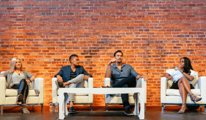 Two men sitting on white chairs against a brick wall, engaged in conversation.