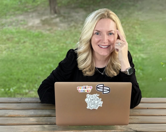 Smiling woman working on a laptop outdoors at a wooden table.